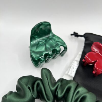 Sodrios žalios spalvos plaukų segtukas žnyplės baltame fone šalia juodo maišelio ir žalios plaukų gumytės