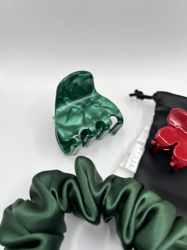 Sodrios žalios spalvos plaukų segtukas žnyplės baltame fone šalia juodo maišelio ir žalios plaukų gumytės