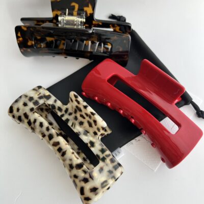 Trijų skirtingų spalvų didesni plaukų segtukai žnyplės baltame fone ant juodo medžiaginio maišelio. Vienas segtukas raudonas, kitas šviesus leopardinis ir kitas tamsios kavos spalvos