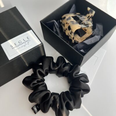 Plaukų aksesuarų rinkinys. Plaukų segtukas žnyplės juodoje dėžutėje ir šalia baltame fone juoda šilkinė plaukų gumytė