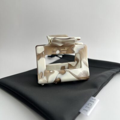 Baltos spalvos su rudais raštais kvadratinis plaukų segtukas žnyplės. segtukas padėtas ant juodo maišelio baltame fone