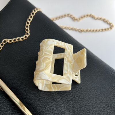 Kvadratinės formos šviesių gelsvų ir baltų spalvų plaukų segtukas žnyplės ant juodos delninukės