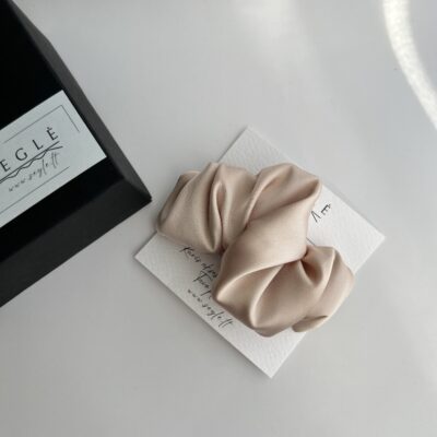 Šviesios kreminės pudros spalvos plaukų segtukas. Segtukas pagamintas iš dirbtinio šilko, savo dizainu panašus į krusaną. segtukas baltame fone šalia juodos dėžutės