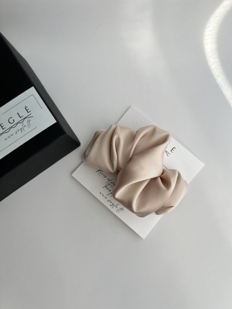 Šviesios kreminės pudros spalvos plaukų segtukas. Segtukas pagamintas iš dirbtinio šilko, savo dizainu panašus į krusaną. segtukas baltame fone šalia juodos dėžutės