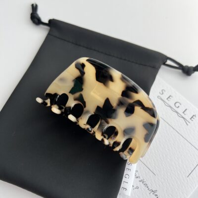 Plaukų segtukas žnyplės šviesios migdolinės spalvos su juodomis dėmelėmis. Segtukas baltame fone ant juodo maišelio