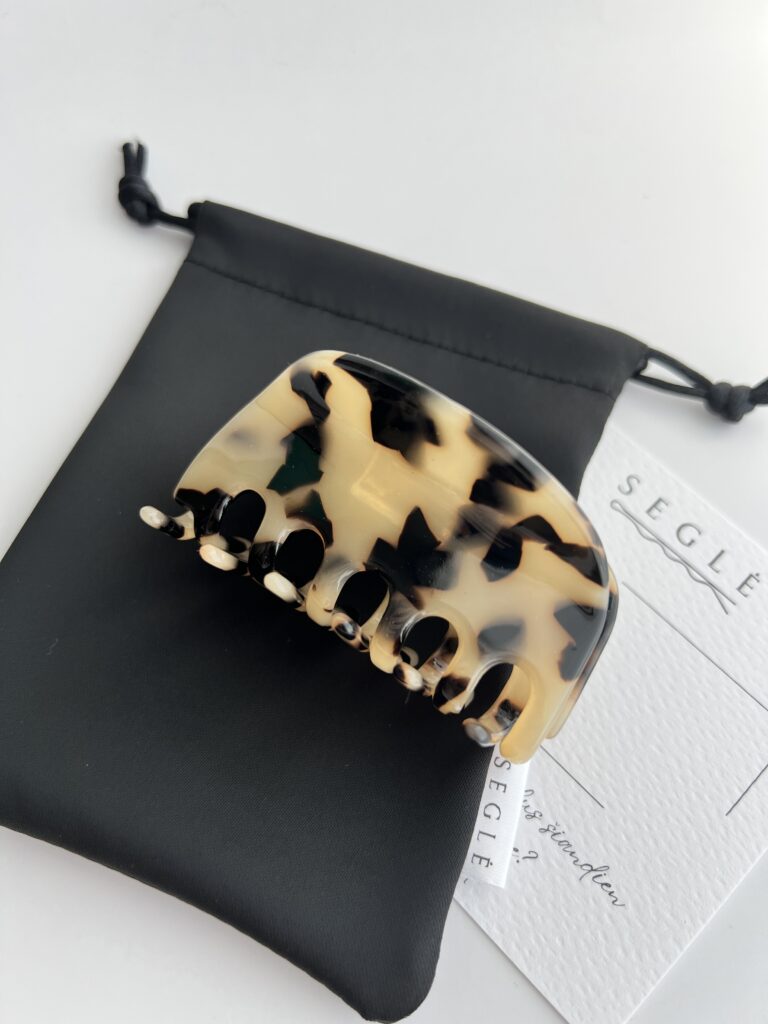 Plaukų segtukas žnyplės šviesios migdolinės spalvos su juodomis dėmelėmis. Segtukas baltame fone ant juodo maišelio