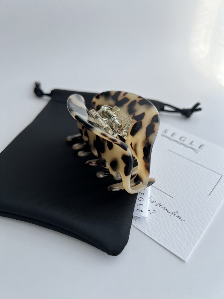 Migdolinės spalvos su juodomis dėmelėmis plaukų segtukas žnyplės baltame fone ant juodo maišelio