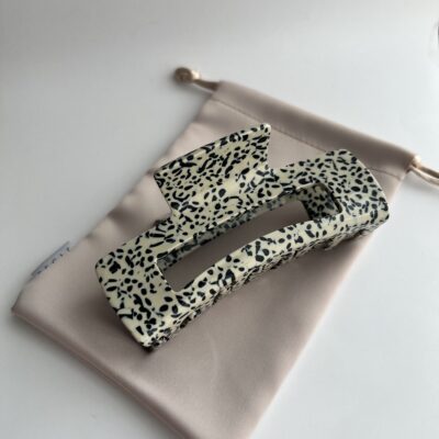 Didelis plaukų segtukas kreminės spalvos su juodais taškeliais. Segtukas baltame fone ant rudo maišelio