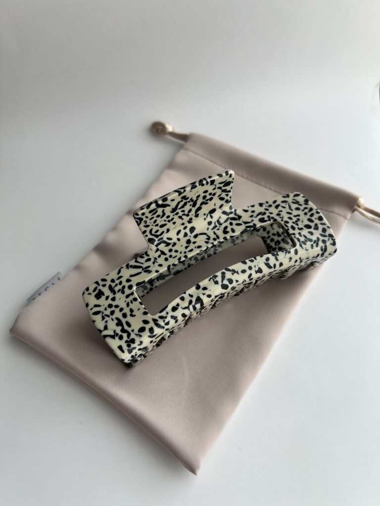 Didelis plaukų segtukas kreminės spalvos su juodais taškeliais. Segtukas baltame fone ant rudo maišelio