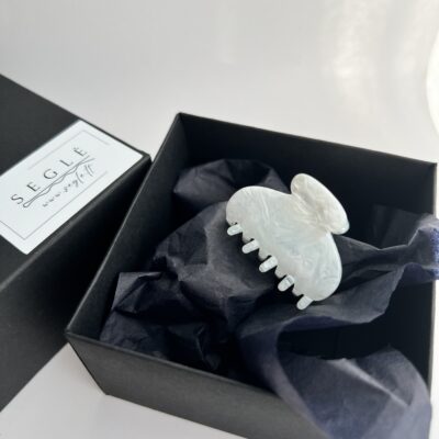 Nedidelis pilkai melsvos spalvos plaukų segtukas žnyplės juodoje dėžutėje. Dėžutė baltame fone