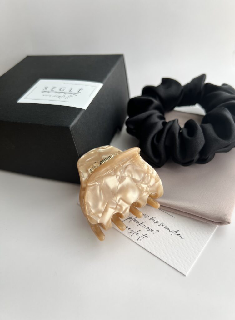 Juodos spalvos šilkinė plaukų gumytė ir karamelinės spalvos plaukų segtukas žnyplės baltame fone šalia juodos dėžutės