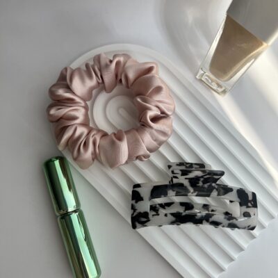 Natūralaus šilko rausvos spalvos šilkinė plaukų gumytė baltame fone šalia plaukų segtuko, pudros buteliuko ir žalio buteliuko