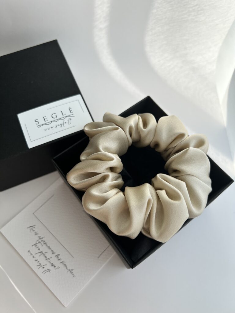 Šampaninės spalvos šilkinė plaukų gumytė juodoje dėžutėje. Dėžutė baltame fone