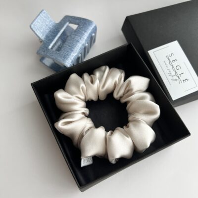 Perlo spalvos šilkinė plaukų gumytė juodoje dėžutėje baltame fone. Šalia dėžutės melsvas plaukų segtukas