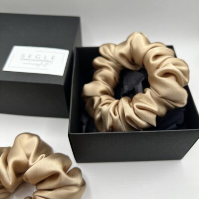 Ryškios aukso spalvos šilkinė plaukų gumytė juodoje dėžutėje, fone dėžutės dangtelis ir kita šilkinė plaukų gumytė