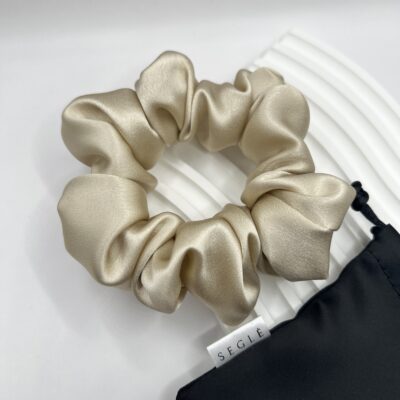 Šampaninės spalvos šilkinė plaukų gumytė baltame fone šalia juodo maišelio