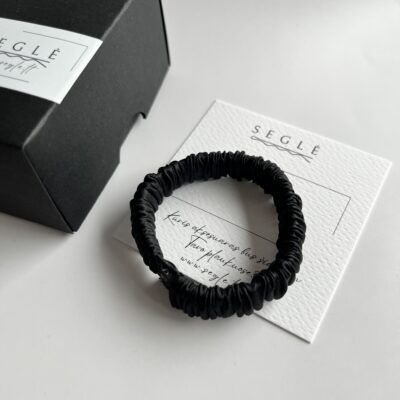 Juodos spalvos siaura šilkinė plaukų gumytė baltame fone šalia juodos dėžutės