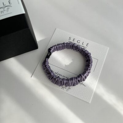 Siaura levandų spalvos šilkinė plaukų gumytė baltame fone šalia juodos dėžutės