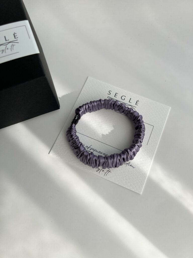 Siaura levandų spalvos šilkinė plaukų gumytė baltame fone šalia juodos dėžutės