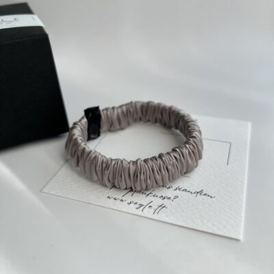 Pilkos spalvos siaura šilkinė plaukų gumytė baltame fone šalia juodos dėžutės