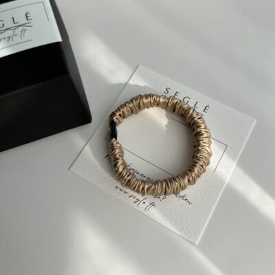 Siaura šilkinė plaukų gumytė. Plaukų gumytė, kreminės, auksinės spalvos. Gumytė baltame fone šalia juodos dėžutės