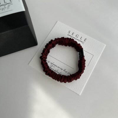 Siaura, bordo spalvos šilkinė plaukų gumytė baltame fone, šalia juodos dėžutės