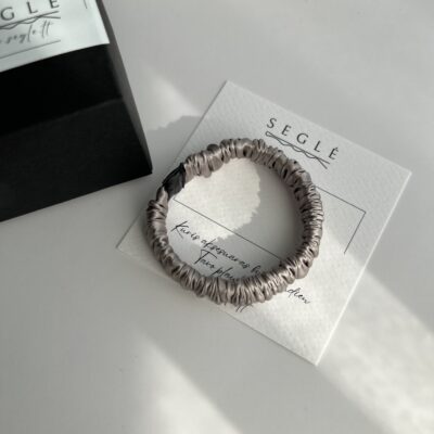 Pilkos spalvos siaura šilkinė plaukų gumytė baltame fone šalia juodos dėžutės