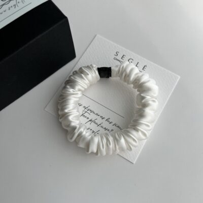 Balta siaura šilkinė plaukų gumytė baltame fone šalia juodos dėžutės