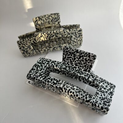 Du dideli plaukų segtukai žnyplės baltame fone. Vienas segtukas kreminės spalvos su juodais taškeliais, kitas baltos spalvos su juodais taškeliais.