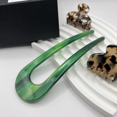 U formos smeigtukas plaukams. Smeigtukas žalios spalvos baltame fone šalia juodos dėžutės ir dviejų nedidelių plaukų segtukų