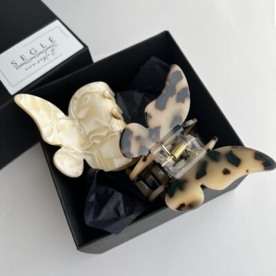 Du drugelio formos plaukų segtukai juodoje dėžutėje. Vienas segtukas baltai gelsvos spalvos, o kitas šviesiai rudos spalvos su juodomis dėmelėmis