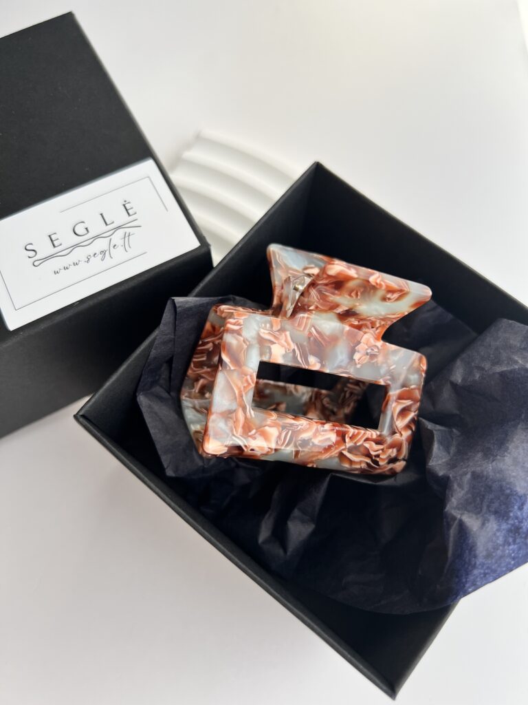 Kvadratinis plaukų segtukas žnyplės juodoje dėžutėje. Segtukas pilkai melsvos spalvos su rudais ornamentais