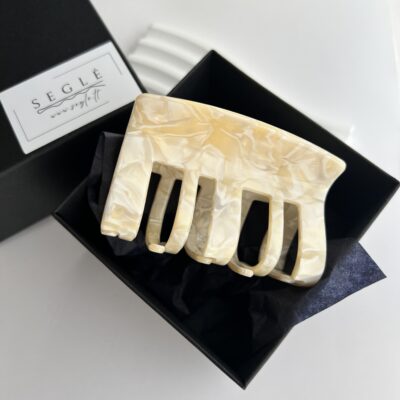 Didelis plaukų segtukas žnyplės juodoje dėžutėje. Segtuko paviršius atrodo lyg išlietas baltas šokoladas