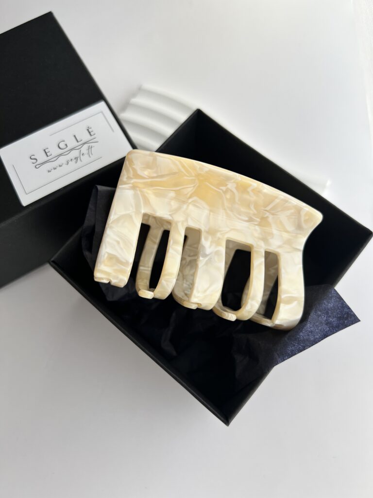 Didelis plaukų segtukas žnyplės juodoje dėžutėje. Segtuko paviršius atrodo lyg išlietas baltas šokoladas