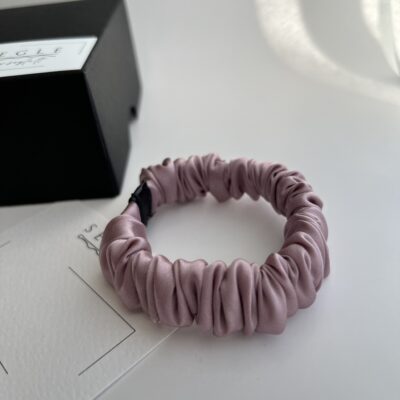 Nedidelė šilkinė plaukų gumytė baltame fone šalia juodos dėžutės. Gumytė švelnios rausvos spalvos