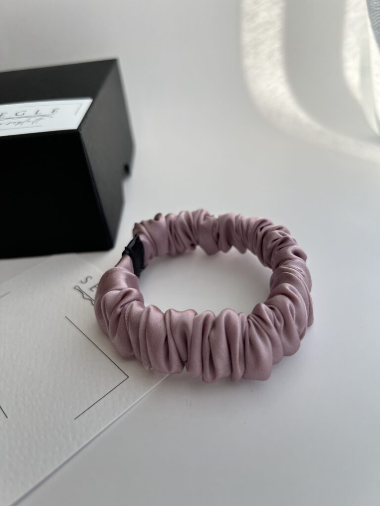 Nedidelė šilkinė plaukų gumytė baltame fone šalia juodos dėžutės. Gumytė švelnios rausvos spalvos