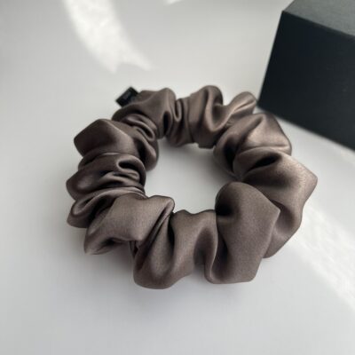 Natūralaus šilko rusvai pilkos spalvos plaukų gumytė baltame fone šalia juodos dėžutės