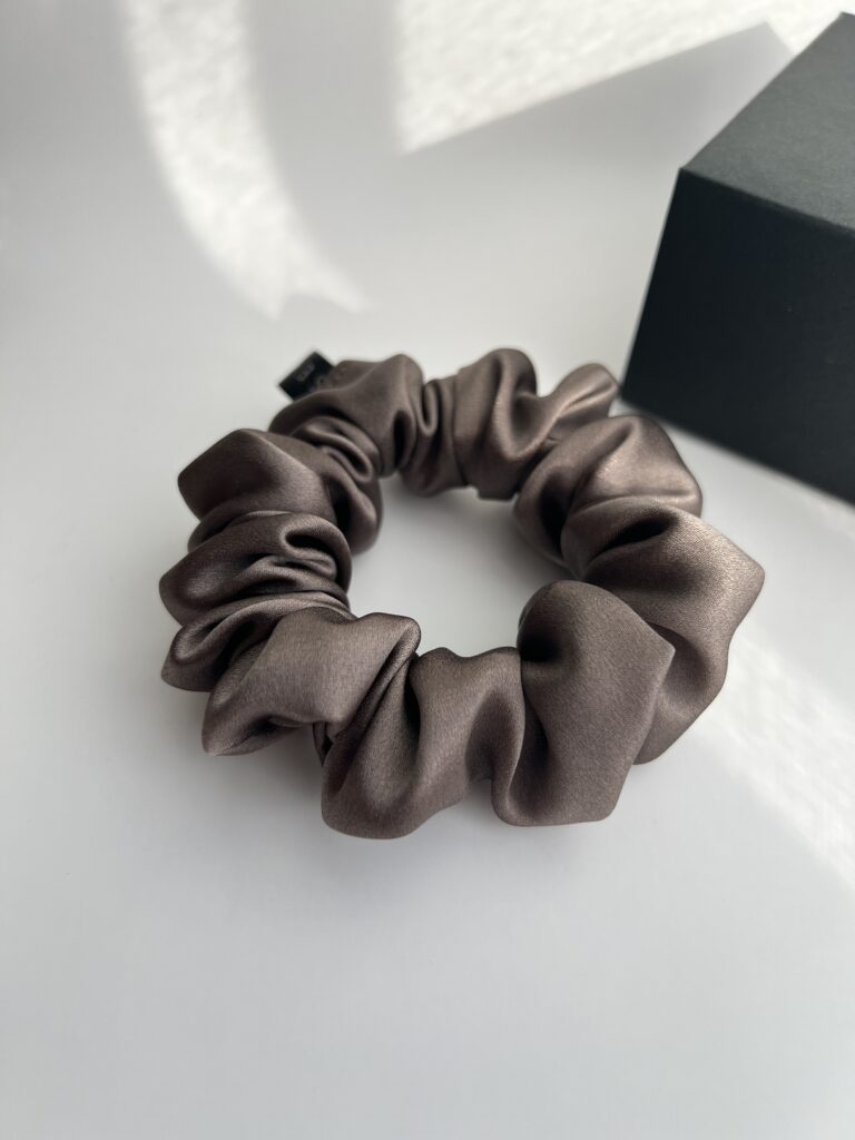 Natūralaus šilko rusvai pilkos spalvos plaukų gumytė baltame fone šalia juodos dėžutės