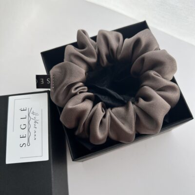 Natūralaus šilko rusvai pilkos spalvos šilkinė plaukų gumytė juodoje dėžutėje. Dėžutė baltame fone