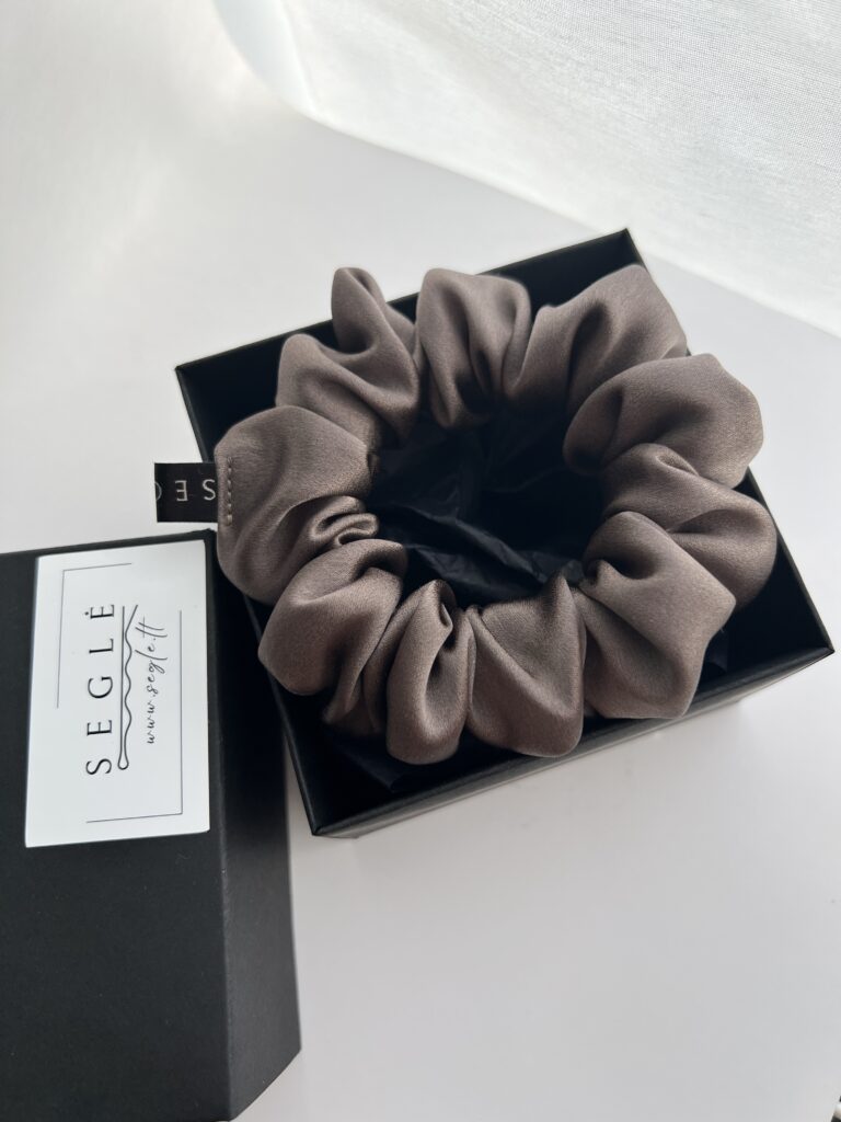 Natūralaus šilko rusvai pilkos spalvos šilkinė plaukų gumytė juodoje dėžutėje. Dėžutė baltame fone