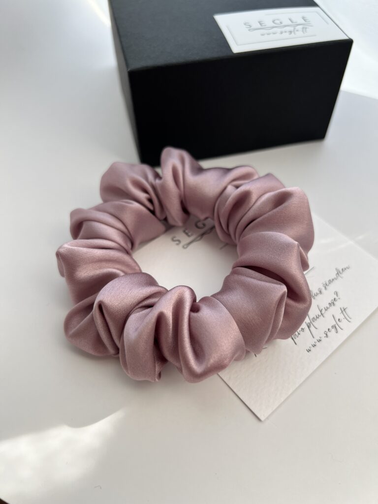 Natūralaus šilko šaltos rausvos spalvos šilkinė plaukų gumytė baltame fone šalia juodos dėžutės