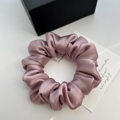 Natūralaus šilko šaltos rausvos spalvos plaukų gumytė. Gumytė baltame fone šalia juodos dėžutės