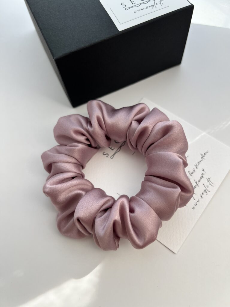 Natūralaus šilko šaltos rausvos spalvos plaukų gumytė. Gumytė baltame fone šalia juodos dėžutės