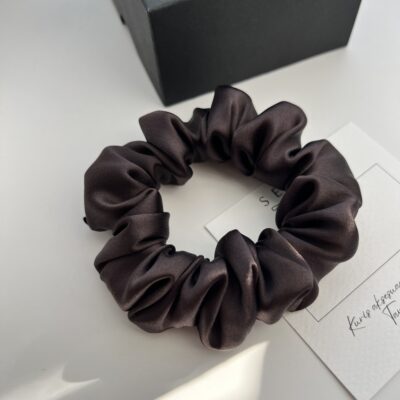 Juodojo šokolado spalvos šilkinė plaukų gumytė baltame fone šalia juodos dėžutės